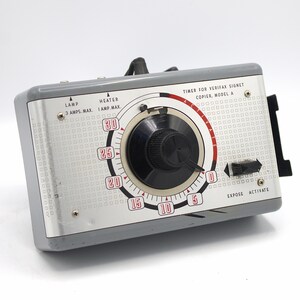 May include: A gray and silver timer for a Verifax Signet copier, Model A. The timer has a large black dial with white and red markings, and a black handle. The timer has the words "EXPOSE ACTIVATE".