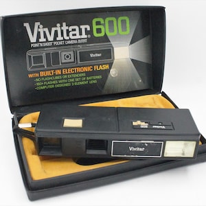 May include: A black Vivitar 600 point and shoot pocket camera outfit with a built-in electronic flash. The camera is in its original box, which has a yellow interior.
