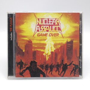 May include: A CD case with a yellow, orange, and red apocalyptic cityscape background. The band Nuclear Assault is printed in black with the album title "Game Over" printed below. The text "includes THE PLAGUE" is printed on the left side of the case.