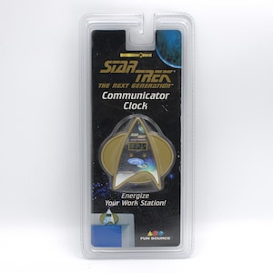 May include: A Star Trek: The Next Generation Communicator Clock in its original packaging. The clock is shaped like the Starfleet insignia and displays the time. The packaging includes the text "Energize Your Work Station!"