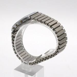 May include: A silver-toned digital wristwatch featuring a rectangular case and a flexible, expandable metal link bracelet. The side of the watch case displays two small circular buttons, and the metallic band consists of numerous rectangular segments.
