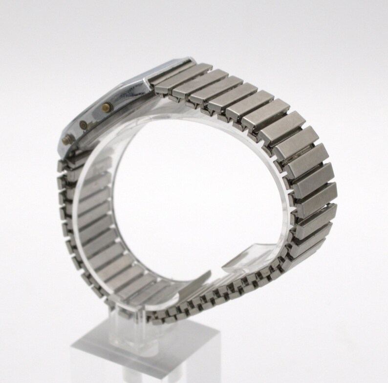 May include: A silver-toned digital wristwatch featuring a rectangular case and a flexible, expandable metal link bracelet. The side of the watch case displays two small circular buttons, and the metallic band consists of numerous rectangular segments.
