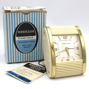 May include: A vintage Westclox travel alarm clock with a cream-colored body and gold-tone accents. The clock has a square face with black numerals and hands. The original box is striped blue and white. The clock is approximately 4 inches wide.