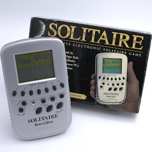 May include: A silver Solitaire Excalibur electronic handheld game with a small screen and multiple buttons. The game is displayed next to its original box, which reads "IVE ELECTRONIC SOLITAIRE GAME."