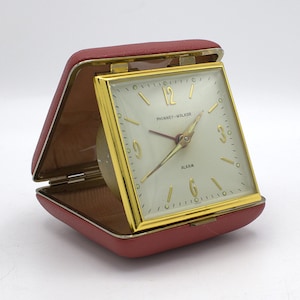 May include: A vintage Phinney-Walker travel alarm clock in a red leatherette case. The clock face is square with gold-tone accents, a cream-colored background, and the word "ALARM" printed on the face.