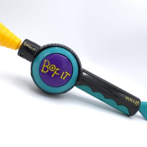May include: A handheld electronic game with a black handle and teal accents. The game features a yellow cone, a purple and teal circular button with the text "Bop It", and the text "Twist It" and "Pull It".