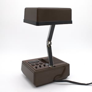 May include: A brown desk lamp with a rectangular head and a black base. The lamp features a metallic, adjustable arm with a central hinge. The base includes a digital display, several black control buttons, and a speaker grille. A black power cord extends from the back of the unit.