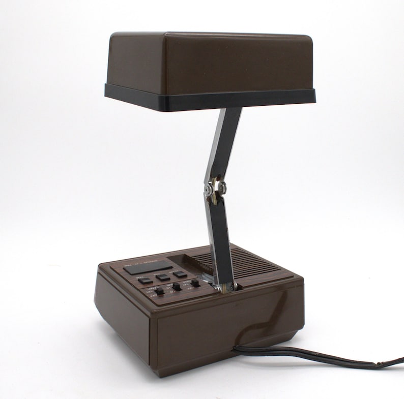 May include: A brown desk lamp with a rectangular head and a black base. The lamp features a metallic, adjustable arm with a central hinge. The base includes a digital display, several black control buttons, and a speaker grille. A black power cord extends from the back of the unit.