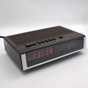 May include: A vintage Lloyds AM/FM electronic clock radio with a brown wood-grain top and black casing. The digital display shows the time in red. The radio has several buttons and a tuning dial.