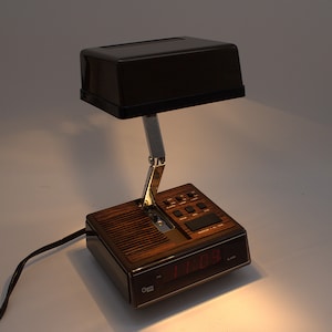 May include: A Cosmo Time alarm clock lamp featuring a dark brown rectangular shade, a chrome adjustable arm, and a base with a wood grain finish. The base includes a digital display with red numbers, control buttons, and a speaker grille.