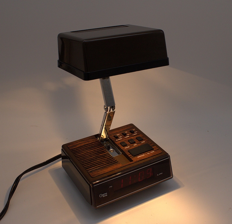 May include: A Cosmo Time alarm clock lamp featuring a dark brown rectangular shade, a chrome adjustable arm, and a base with a wood grain finish. The base includes a digital display with red numbers, control buttons, and a speaker grille.