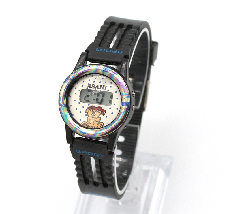 May include: Black Asahi Sport watch with a white face featuring a cartoon lion and the time 2:00. The watch has a black band and a colorful iridescent bezel. The word "SPORT" is printed in blue.