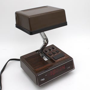 May include: A brown Cosmo Time digital alarm clock with a hinged, adjustable lamp head. The base features a wood-grain panel, a speaker grille, and a digital display showing the time in red. Control buttons for alarm, lamp, hour, and minute are present.