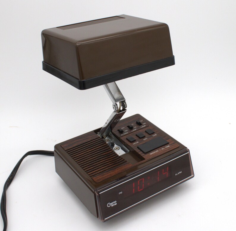 May include: A brown Cosmo Time digital alarm clock with a hinged, adjustable lamp head. The base features a wood-grain panel, a speaker grille, and a digital display showing the time in red. Control buttons for alarm, lamp, hour, and minute are present.