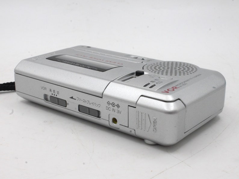 May include: A silver cassette recorder with a red VOR logo and a black button on the top. The recorder has a microphone, a speaker, and a DC in 3V port. The recorder has a few buttons on the side, including a fast forward button and a rewind button.