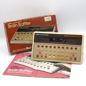 May include: Vintage Mattel Electronics Brain Baffler game. The beige and brown electronic game has multiple colored buttons and a display screen. The original box and instruction manual are also visible.