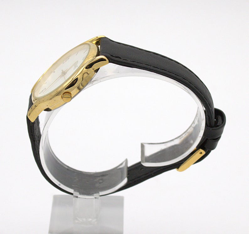 May include: A gold-toned wristwatch with a white face and black leather band. The watch has a classic design with a round face and a buckle closure. The watch is displayed on a clear stand against a white background.