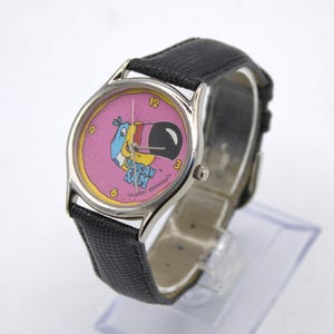 May include: A vintage wristwatch featuring the cartoon character Toucan Sam on a pink dial. The watch has a silver-tone case, black leather band, and displays the time with black numerals. The watch is a collectible item.