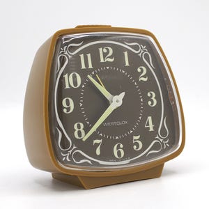 May include: A vintage brown Westclox alarm clock with a dark brown face and white numerals. The clock has a rectangular shape with rounded edges and a clear plastic cover. The hands and hour markers glow in the dark. The words "WESTCLOX" and "MADE IN USA" are visible.