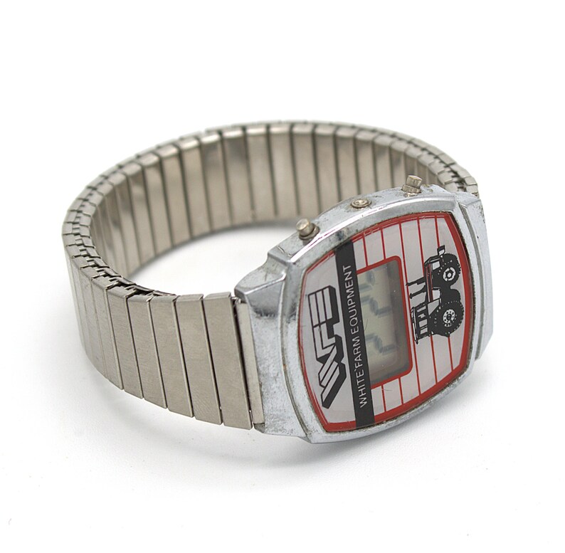 Peut inclure: Montre num&eacute;rique vintage avec un bracelet extensible argent&eacute;. Le cadran est blanc avec un bord rouge et pr&eacute;sente un dessin de tracteur et le texte "WHITE FARM EQUIPMENT". La montre affiche l'heure en num&eacute;rique.