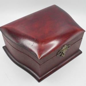 May include: A burgundy-colored wooden box with a curved lid and base. The box features a brass-colored clasp and hinges. The wood grain is visible, adding texture to the design. The box is likely used for storage or display.