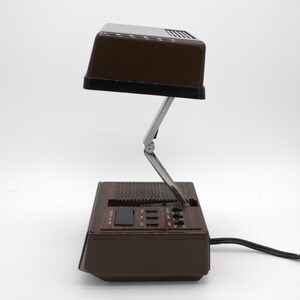 May include: A brown desk lamp with a rectangular head and a folding metal arm. The base features a wood-grain finish, a speaker grille, and a control panel with several black buttons and switches. A black power cord extends from the back of the unit.