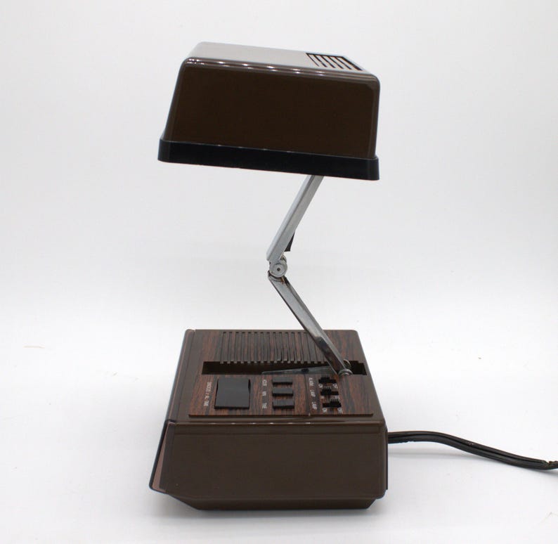 May include: A brown desk lamp with a rectangular head and a folding metal arm. The base features a wood-grain finish, a speaker grille, and a control panel with several black buttons and switches. A black power cord extends from the back of the unit.