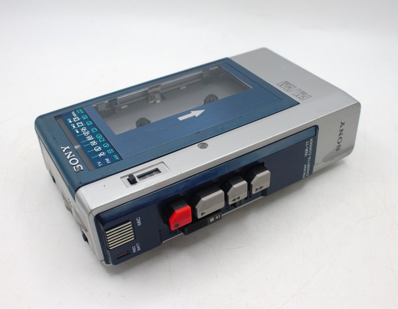 May include: A vintage Sony WM-11 cassette player in silver and blue. The portable audio device features a cassette compartment, control buttons, and a radio dial. The Sony logo is visible on the front and side of the player.
