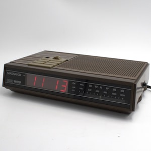May include: A brown Magnavox digital clock radio displaying "11:13" in red digits. The clock has a rectangular design with a speaker grill and control buttons on top. The radio dial includes FM and AM frequencies.