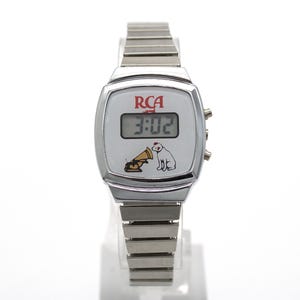 May include: A silver-tone digital wristwatch with a white face featuring the red RCA logo and a graphic of a dog listening to a gramophone. The watch has a rectangular digital display showing 3:02 and a flexible metal link band with a polished finish.