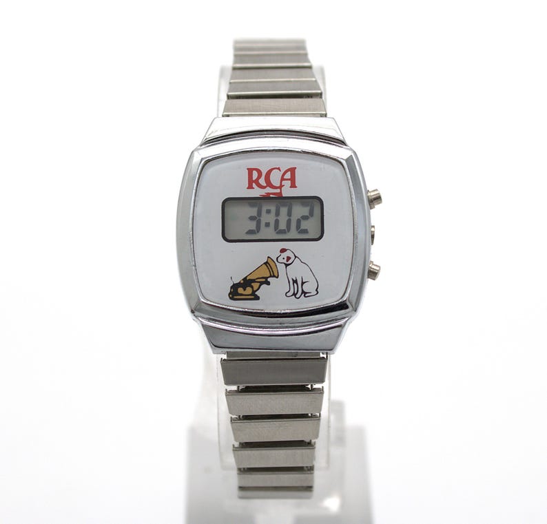 May include: A silver-tone digital wristwatch with a white face featuring the red RCA logo and a graphic of a dog listening to a gramophone. The watch has a rectangular digital display showing 3:02 and a flexible metal link band with a polished finish.