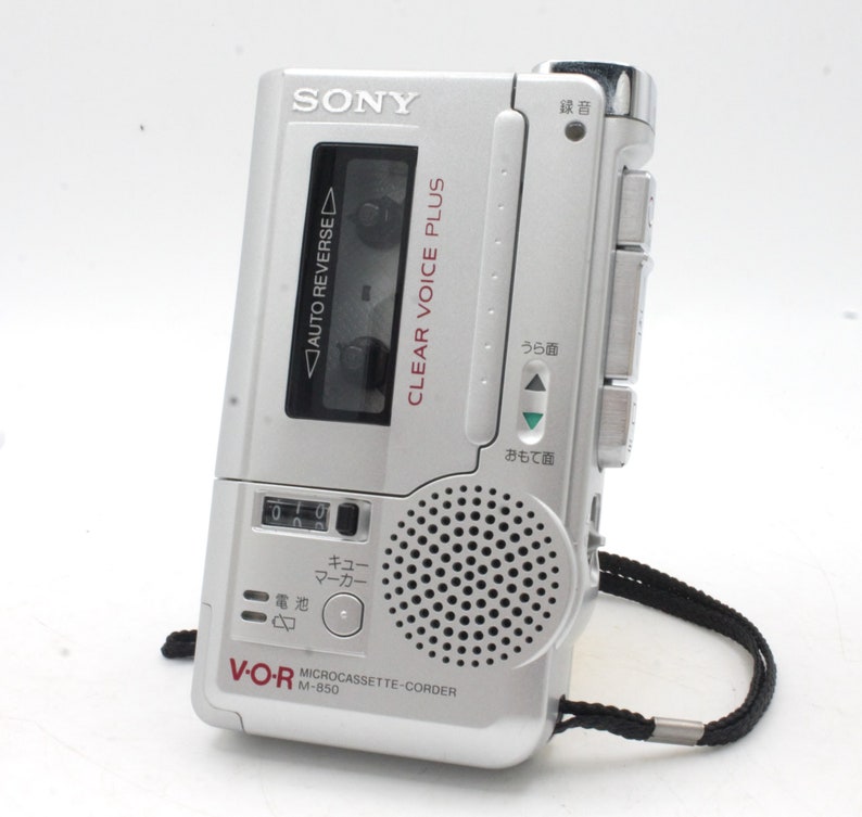 May include: A silver Sony V-O-R microcassette recorder with a black strap. The recorder has a clear voice plus feature and a counter display. The text on the recorder reads "CLEAR VOICE PLUS" and "MICROCASSETTE-CORDER M-850".