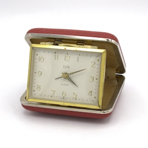 May include: A vintage Elgin travel alarm clock in a red case. The clock face is white with gold-colored hands and a gold-colored frame. The word "ELGIN" is printed on the clock face. The case is open, revealing the clock.
