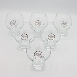 May include: Set of six clear glass shot glasses with a round base. Each glass has a gold and black logo that reads "Mozart Liquore".