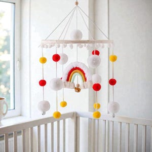 May include: A baby mobile featuring a rainbow, white pom-poms, and red and yellow accents. The mobile is suspended from a wooden ring and hangs above a white crib. The rainbow has a small teddy bear attached.