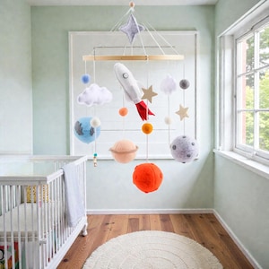 Op de afbeelding: Een baby mobile met een ruimtethema, met een witte raket, planeten, sterren en wolken. De mobile hangt boven een wit babybedje in een kamer met een houten vloer en een rond, wit tapijt. De planeten zijn oranje, perzik en grijs.