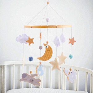 Op de afbeelding: Een houten baby mobile met een lichtbruin houten frame. Het bevat een maan, sterren, wolken, een astronaut en een raket, in zachte pastelkleuren. Het hangt boven een wit babybedje.