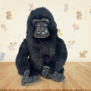 May include: A plush gorilla toy with black fur and a detailed face. The gorilla has a realistic appearance with brown eyes and a leather-like face and hands. The toy is sitting on a wooden surface.