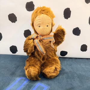 May include: A plush monkey doll with brown fur, a light tan fabric face, and a multi-colored striped ribbon tied around its neck. The doll has a rounded head, small ears, and one arm raised in a waving position, featuring a soft, textured appearance.