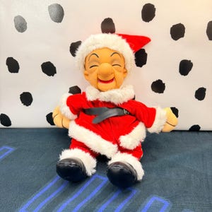 May include: A plush doll featuring a tan face with closed eyes and a wide smile, wearing a red velvet Santa suit with white faux fur trim, a matching Santa hat, a black belt, and black boots. The doll has a soft, rounded nose and ears.