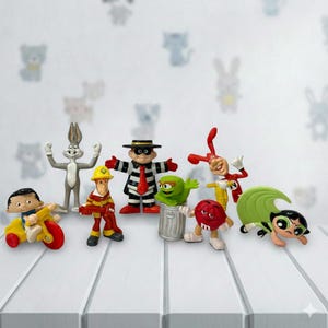 May include: A selection of colourful character figurines, including Bugs Bunny, a firefighter, a Hamburglar, and others. The figurines are arranged on a white surface with a patterned background. The characters are from various cartoons and shows.