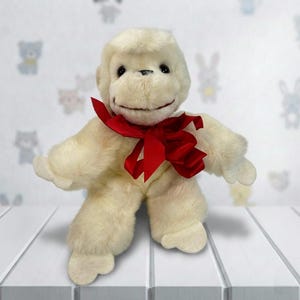 May include: A soft, off-white monkey toy with a red bow. The toy has a smiling face and is placed on a white surface. The background has a pattern of cartoon animals.