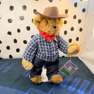 May include: A golden-brown plush teddy bear wearing a tan cowboy hat, a red bandana with a white paisley pattern, a navy blue and white plaid button-down shirt, and dark blue denim jeans. A tag reads The Vermont Teddy Bear Complete Companion.