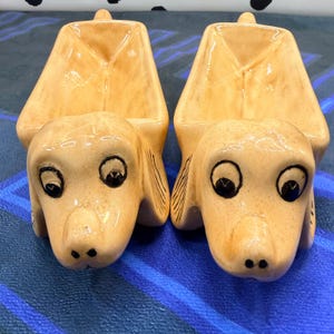 May include: Two matching ceramic dog-shaped planters with a glossy light tan finish. Each piece features a stylised canine head with painted black eyes and nose, and a hollow, open-topped body section designed to hold small items or plants.