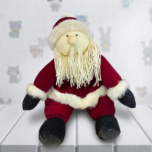 Op de afbeelding: Een pluche kerstmanpop met een rood fluwelen pak, witte imitatiebontafwerking en een bijpassende hoed. De pop heeft een crèmekleurig gezicht, een lange witte baard en zwarte laarzen. Een feestelijke kerstversiering.