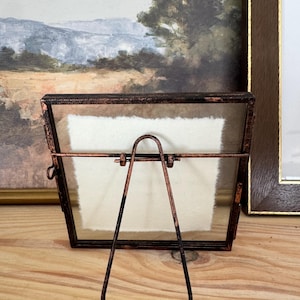 May include: A small, rectangular picture frame with a copper-colored metal frame and a glass front. The frame has a built-in easel stand. The frame is empty, with a white paper insert. The frame is sitting on a wooden surface.