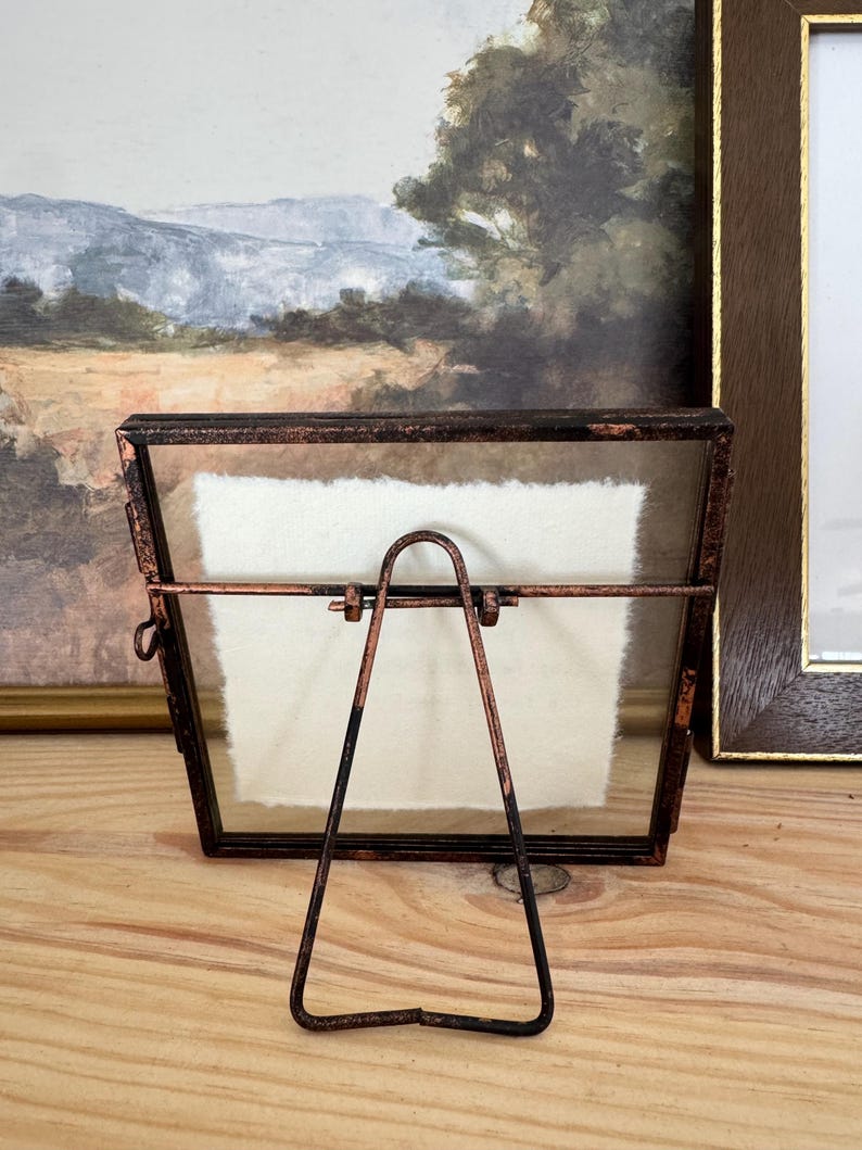 May include: A small, rectangular picture frame with a copper-colored metal frame and a glass front. The frame has a built-in easel stand. The frame is empty, with a white paper insert. The frame is sitting on a wooden surface.