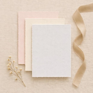 May include: Three textured paper cards in pastel pink, cream, and white, arranged with a beige ribbon and a sprig of dried flowers. The cards have deckled edges, suggesting a handmade or artisanal quality, suitable for stationery or invitations.