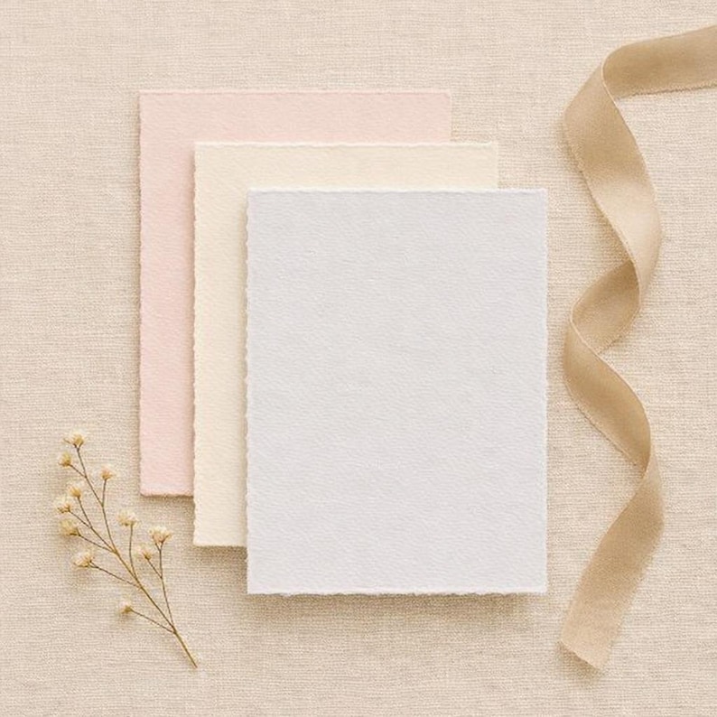 May include: Three textured paper cards in pastel pink, cream, and white, arranged with a beige ribbon and a sprig of dried flowers. The cards have deckled edges, suggesting a handmade or artisanal quality, suitable for stationery or invitations.