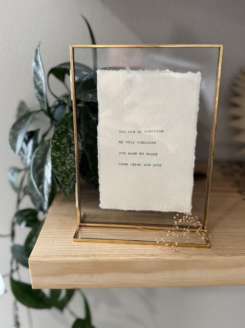 Pu&ograve; includere: Una stampa incorniciata con il testo "You are my sunshine, my only sunshine, you make me happy, when skies are grey." La cornice &egrave; dorata e rettangolare, con un fronte in vetro trasparente. La carta all'interno &egrave; bianco sporco.